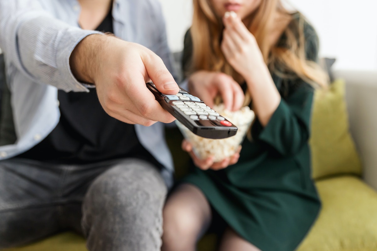 Duas pessoas assistindo algo na televisão. Uma delas aponta o controle para a TV e pega pipoca.