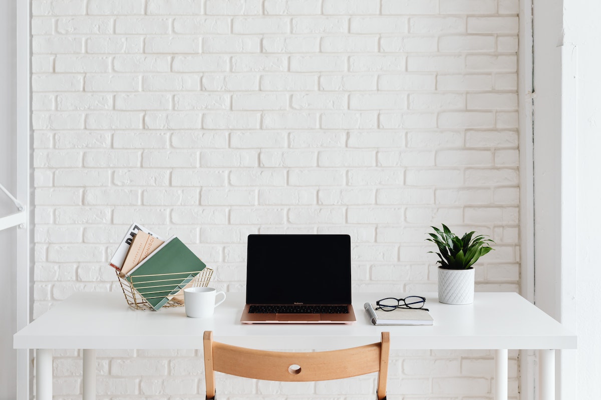 Um botebook em cima de uma mesa de escritório. Ao lado esquerdo há alguns livros enquanto do lado direito tem um pequeno vaso com planta, e um óculos em cima do caderno