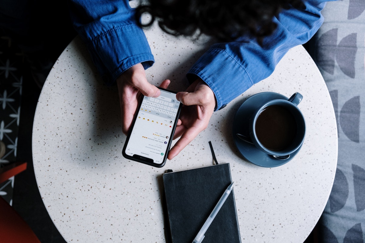 Vista aérea de mulher cobrando seu cliente pelo WhatsApp. Na mesa tem uma agenda fechada com uma canta em cima, e uma xícara de café ao lado