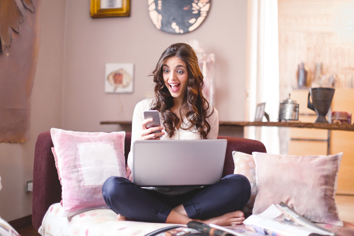 Uma mulher sentada na cama. Em seu colo está seu notebook e no smartphone em sua mão ela está vendo uma lista de Celular Barato 2023