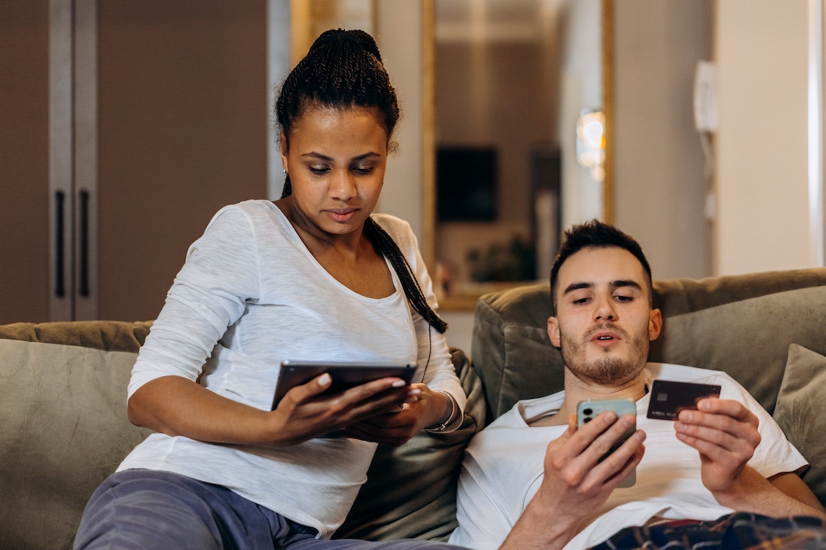 Casal sentado no sofá. A mulher olha o tablet enquanto o marido confere informações no cartão e no aplicativo de celular