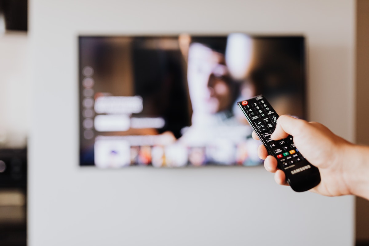 Foco em uma mão segurando o controle da televisão. Ao fundo, a tv aparece desfocada