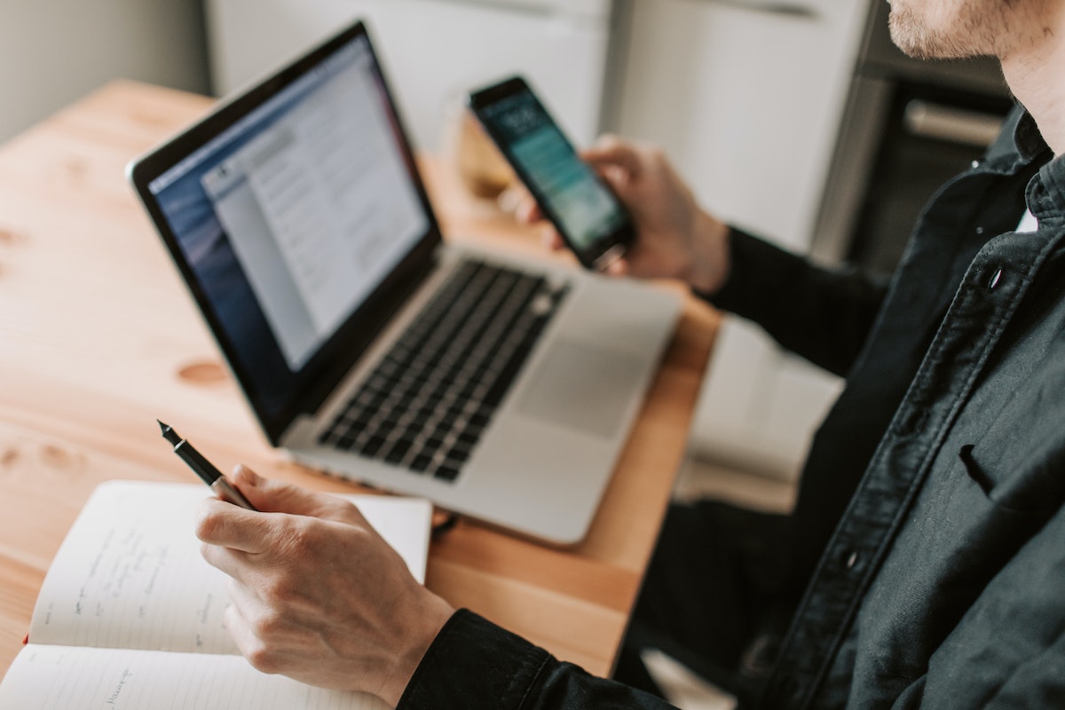 Pessoa mexendo no celular enquanto observa o seu notebook e anota algo em seu caderno