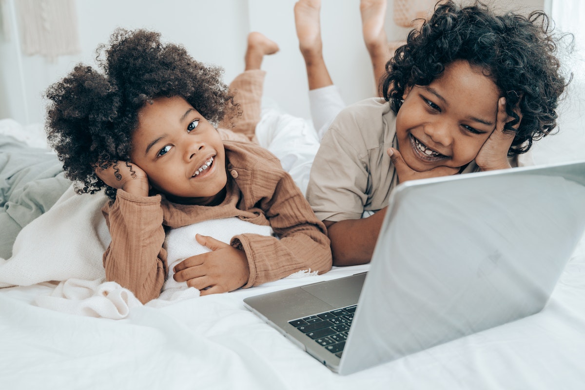 Duas crianças estão deitadas de bruços em frente a um notebook na cama