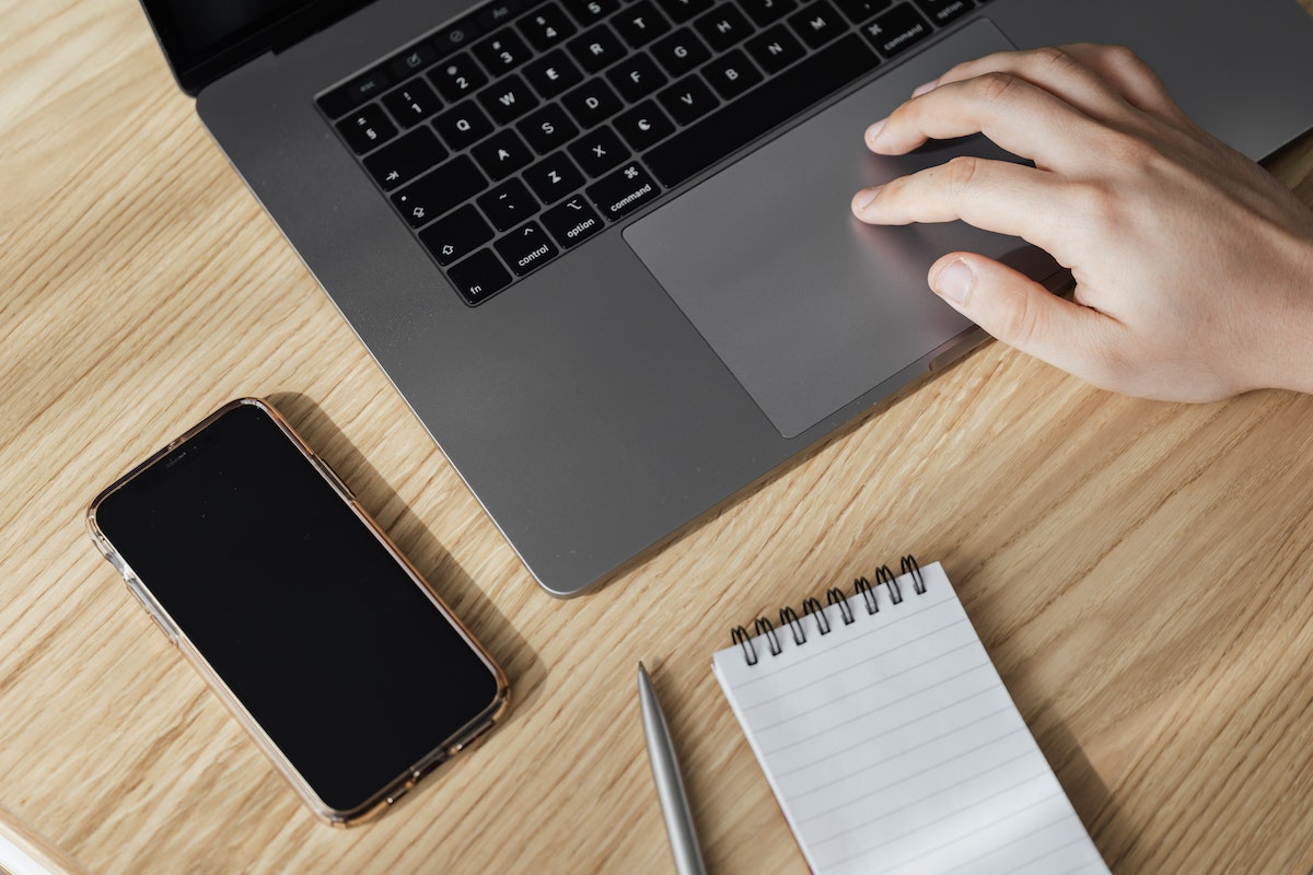 Homem mexendo em um notebook. Na mesa tem um smartphone bloqueado e um bloco de notas com caneta ao lado 