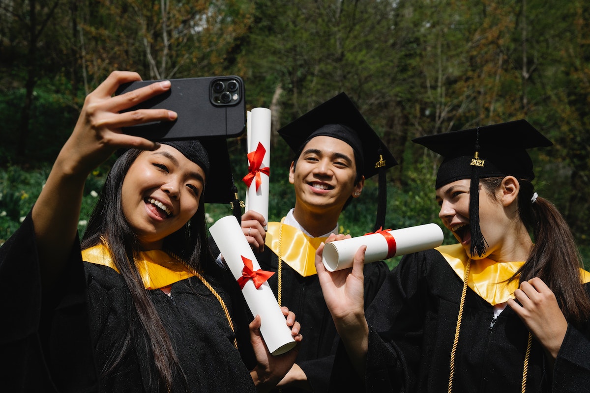 Uma menina tira uma selfie com uma amiga e um amigo. Eles estão de beca e com os diplomas nas mãos