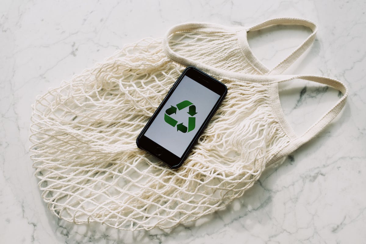 A foto de um celular com a logo de reciclagem na tela. Ele está em cima de uma bolsa de renda e é um celular descontinuado