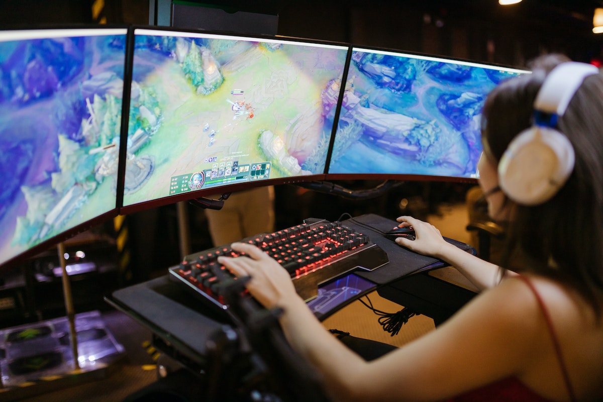 Mulher jogando videogame em dois monitores. Sentada em uma cadeira de escritório, com os dois monitores posicionados à sua frente. Usando headset e segurando um controle de videogame em cada mão.