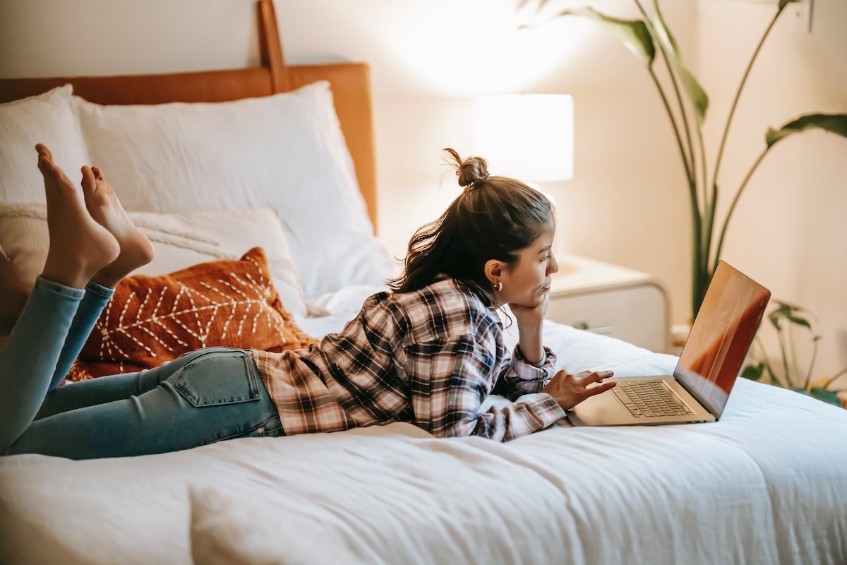 Uma mulher deitada de bruços em sua cama, mexendo em seu notebook