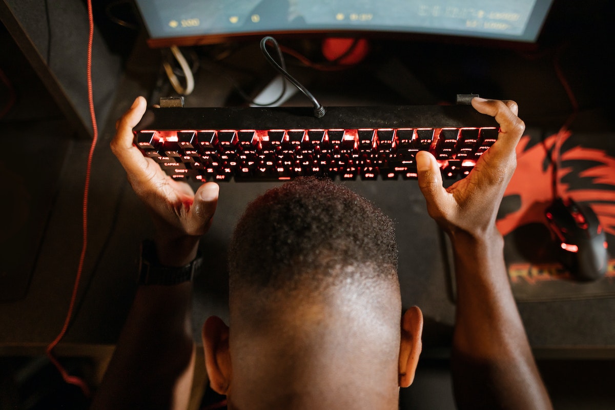 Homem segurando um teclado de computador acima da cabeça, enquanto joga um videogame. Sentado em uma cadeira de escritório, com o computador ligado à sua frente. Usando headset e fazendo uma expressão de concentração.