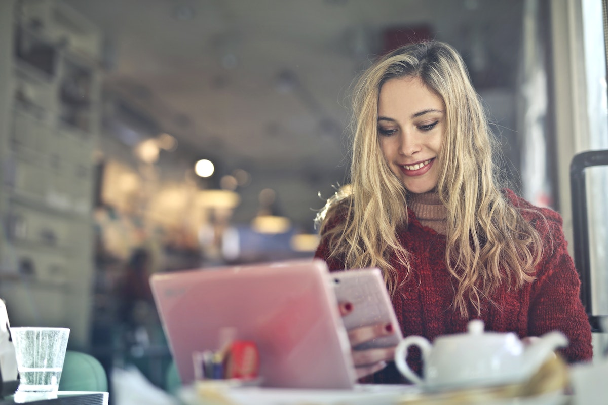 Uma mulher usando um celular na frente do notebook. Ela está sorrindo, em cima da mesa tem diversos itens 