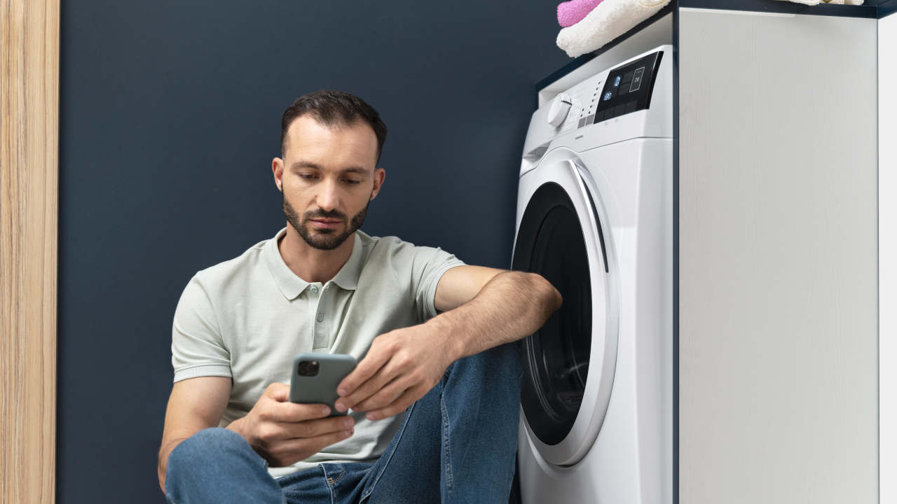 Homem mexendo em celular encostado em máquina de lavar roupas