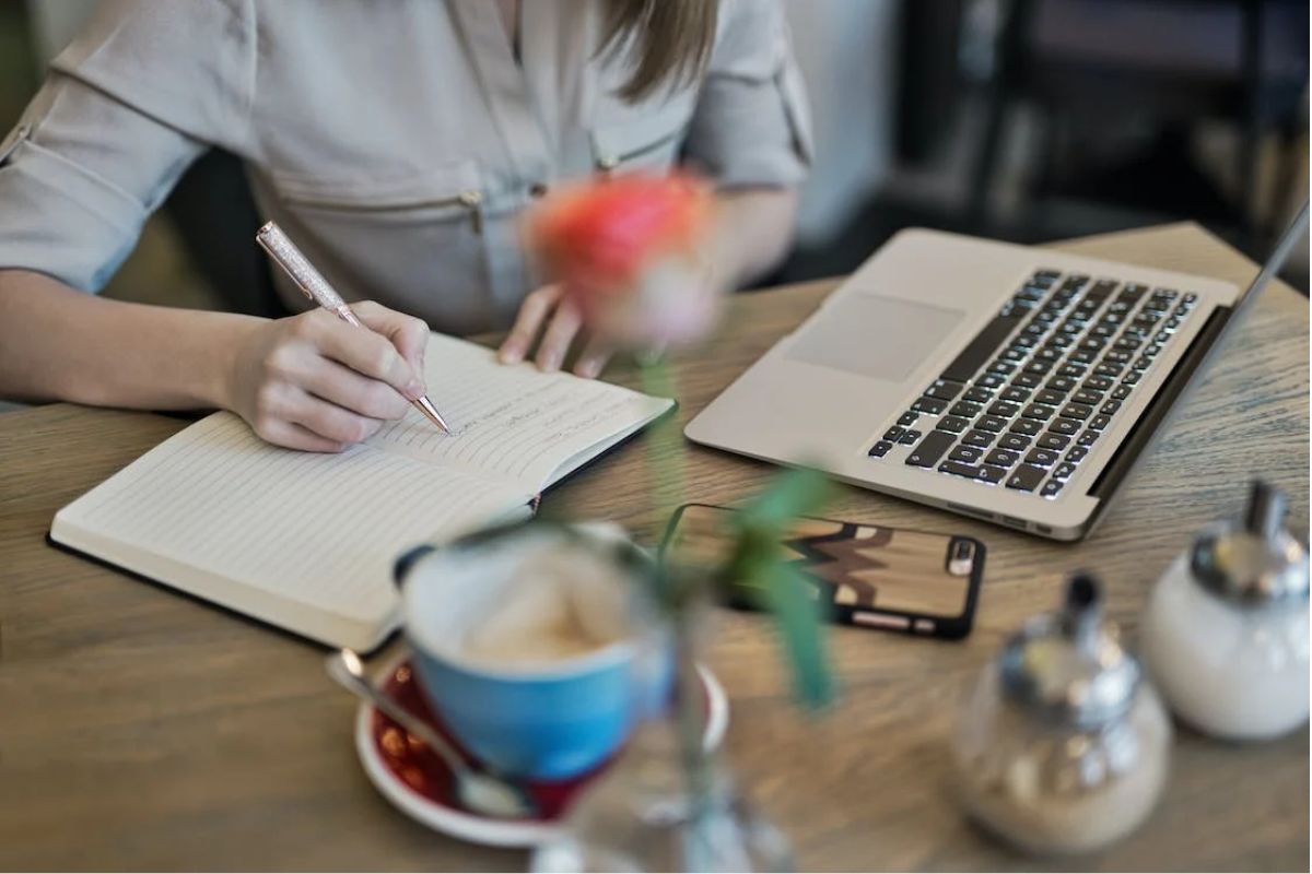 Uma pessoa está na mesa de seu escritório mexendo em um notebook. Ela também faz anotações em um caderno, e há diferentes itens na mesa