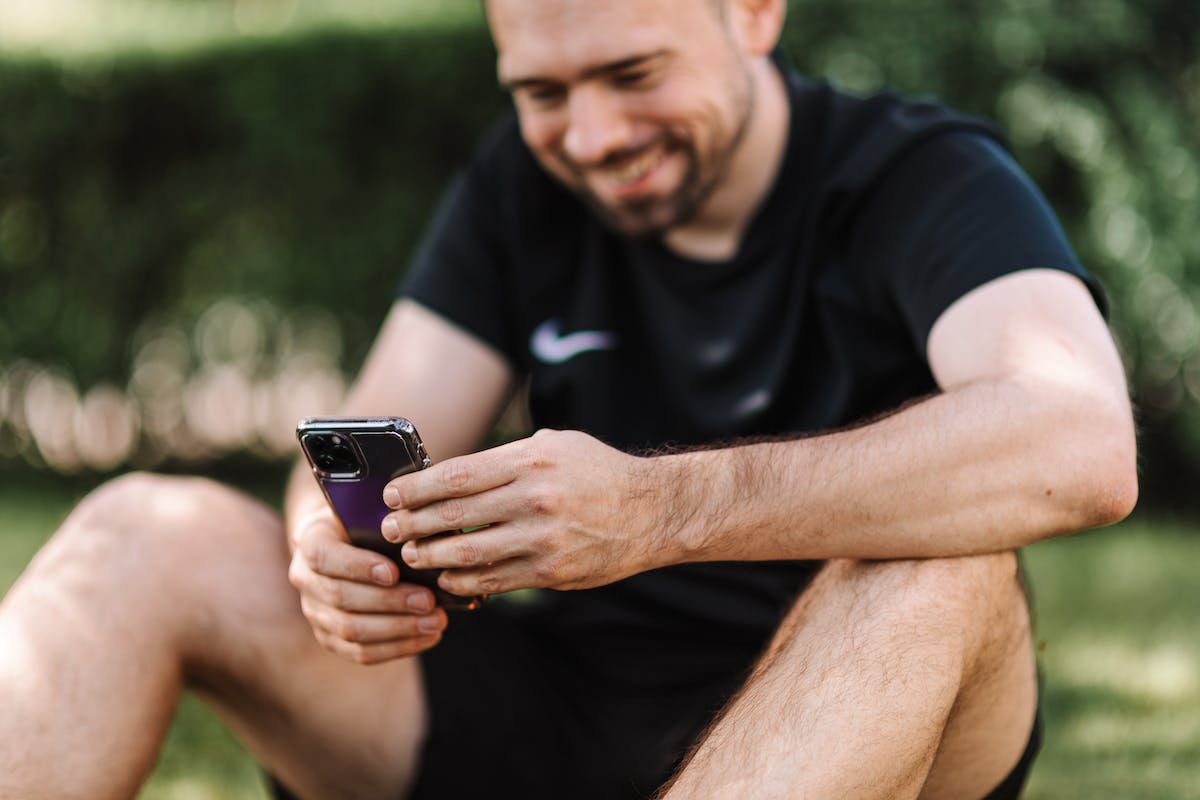 Um rapaz está sentado no campo com o celular na mão usando um Aplicativo de Assistir Jogo ao Vivo