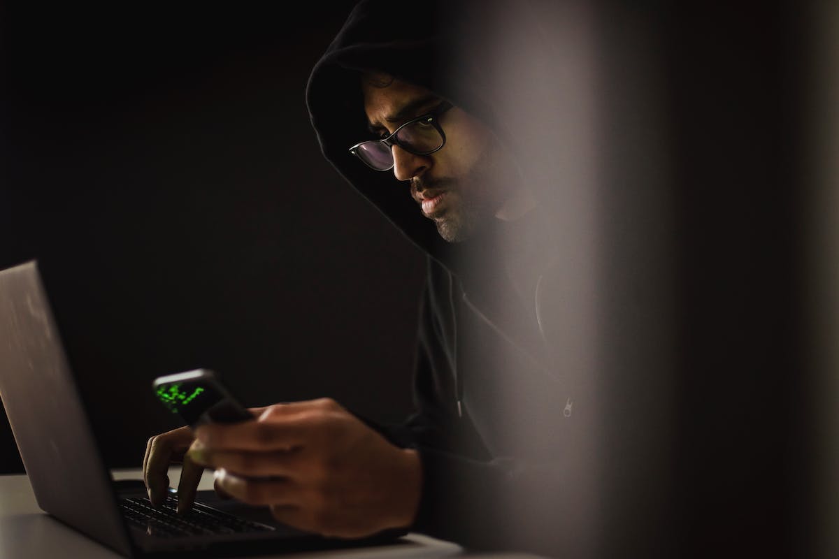 Um espião está de capuz, mexendo no computador com um celular na mão. Ele está em um local escuro e usa óculos
