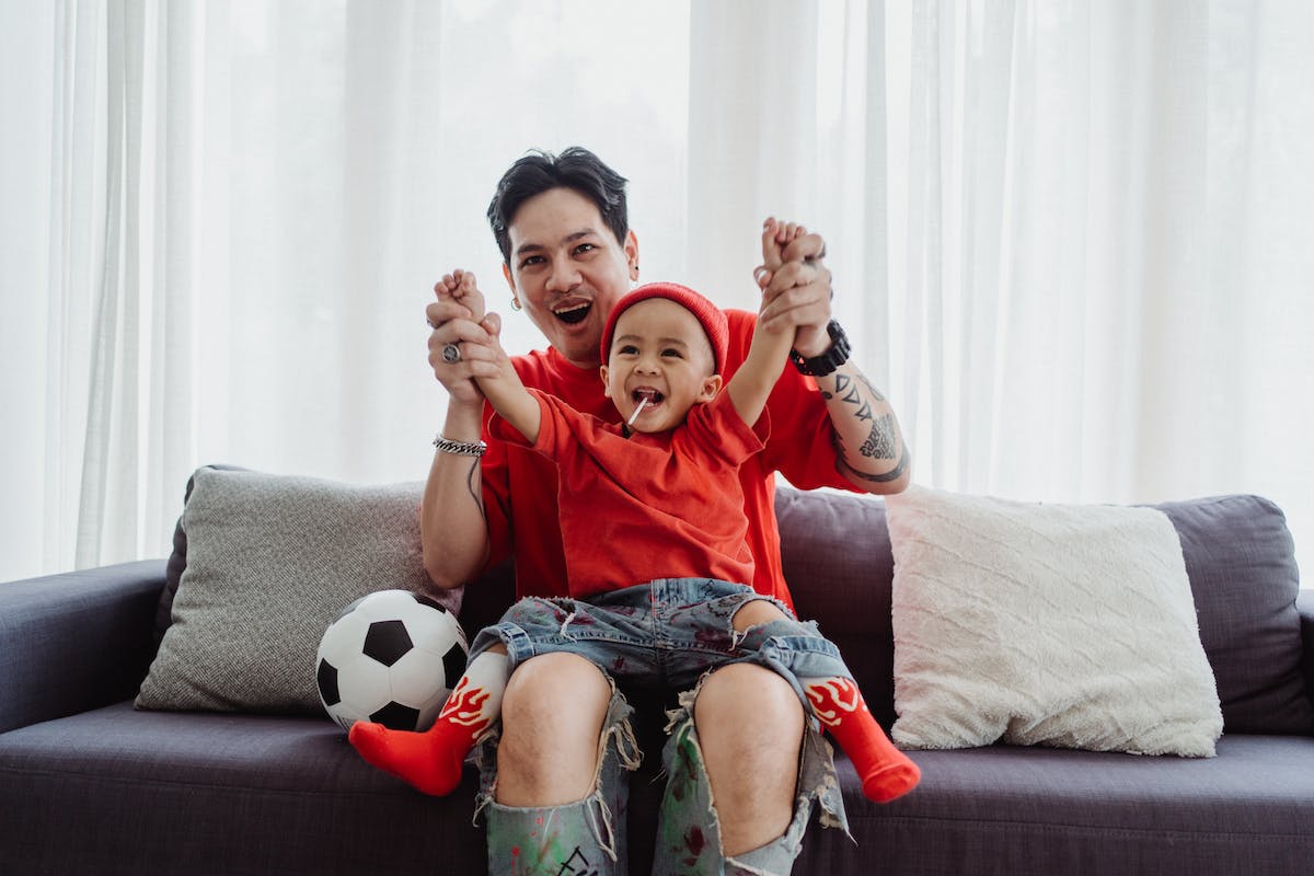 Um pai está com o seu filho no sofá enquanto eles comemoram para o seu time de futebol
