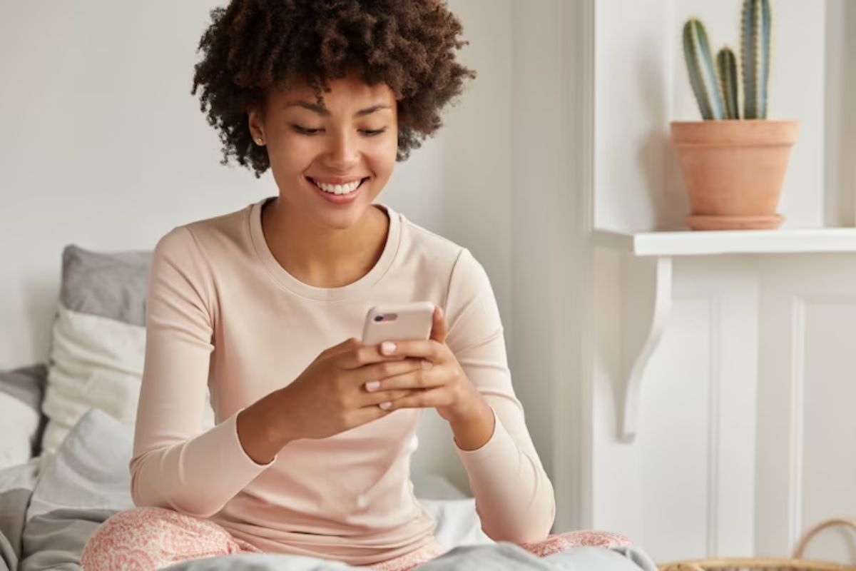 Uma moça está sentada na cama com as duas mãos segurando o celular. Ela sorri enquanto usa um app para ganhar dinheiro com avaliação