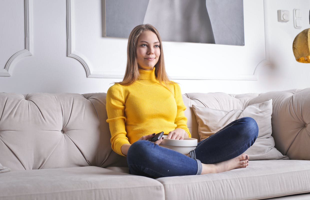 Uma menina de casaco amarelo está sentada vendo filme. Entre suas pernas tem um pote com pipoca