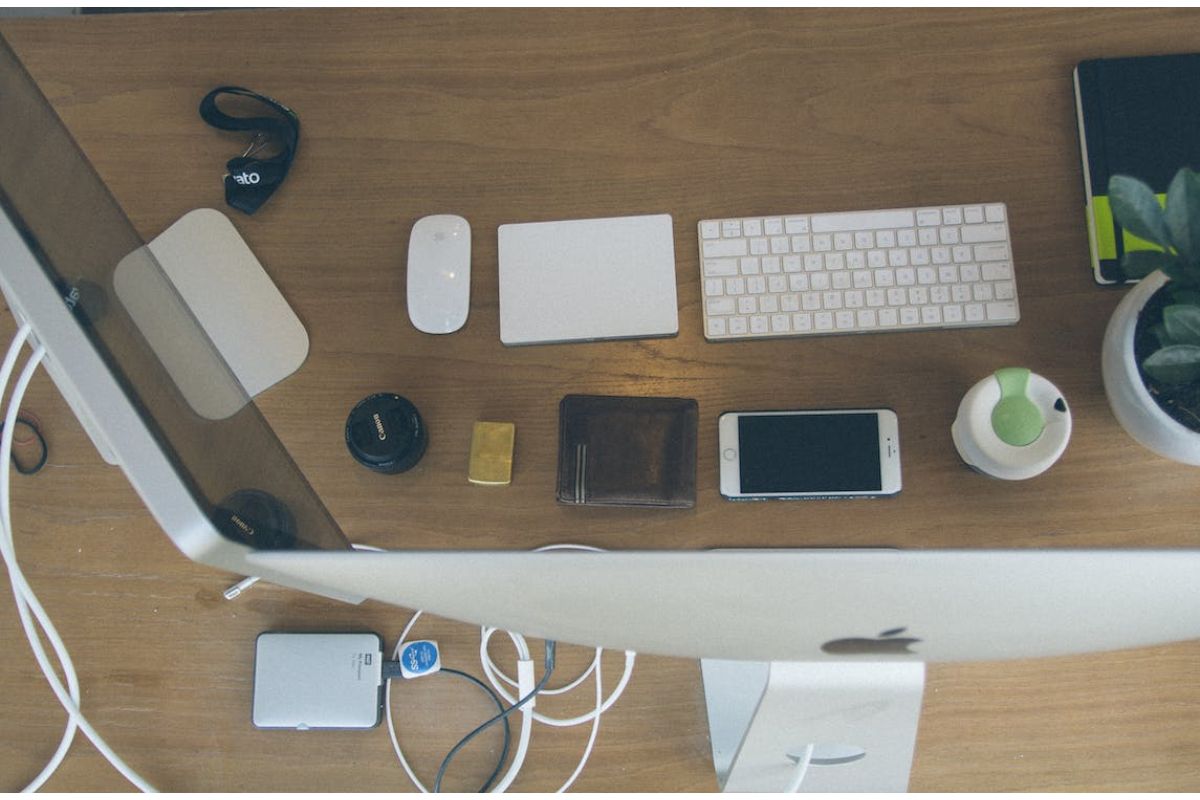 Imagem de acessórios de computador, um Magic Trackpad ou Magic Mouse e teclado em cima da mesa, junto com um celular e carteira