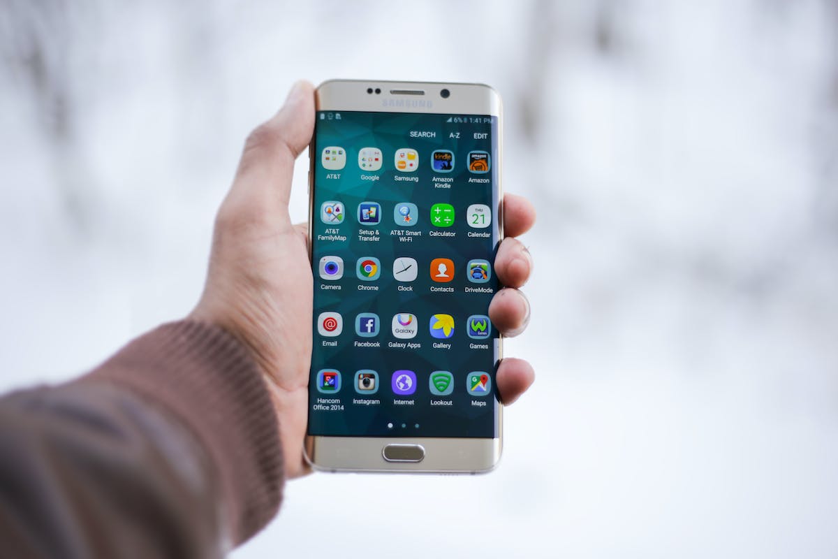 Uma pessoa segura o celular na mão na tela inicial onde é possível ver diferentes aplicativos, incluindo um Aplicativo de Assistir Jogo ao Vivo