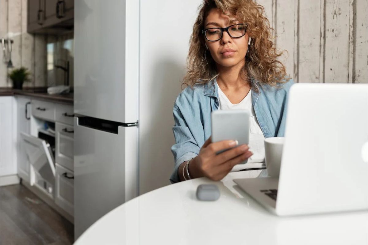 Mulher com um celular na mão. Ela está sentada em uma cadeira em frente ao notebook que está em uma mesa