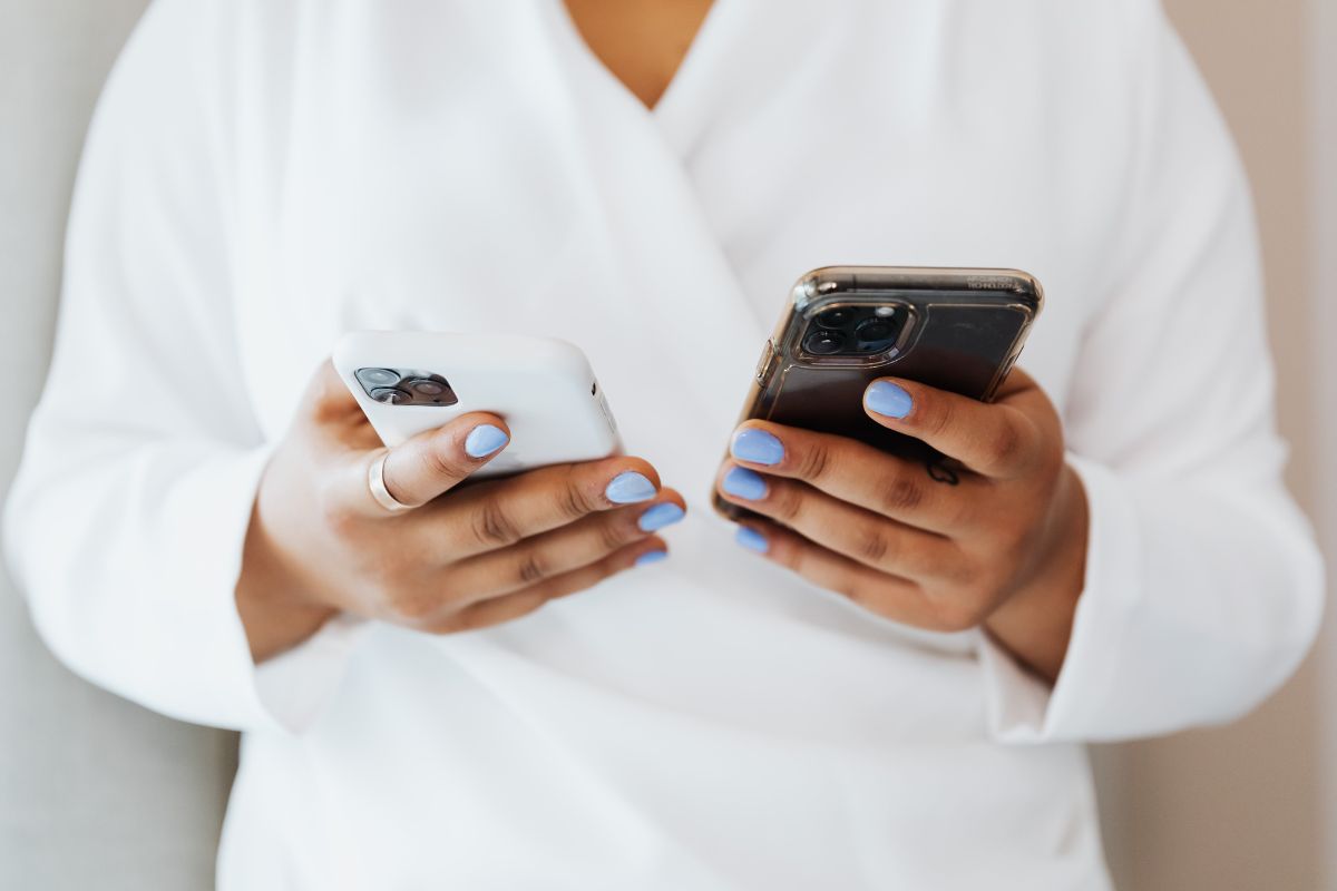 Uma mulher segura dois smartphones em sua mão enquanto decide qual é o melhor. Ela veste uma camisa branca e aparece apenas seu busto