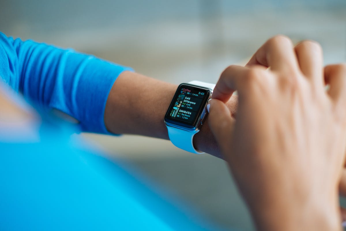 Uma mulher vestindo uma camisa azul e olhando para um smartwatch no pulso. O smartwatch está mostrando a hora e a data. A mulher está de pé vendo se o smartwatch conta frequências cardíacas