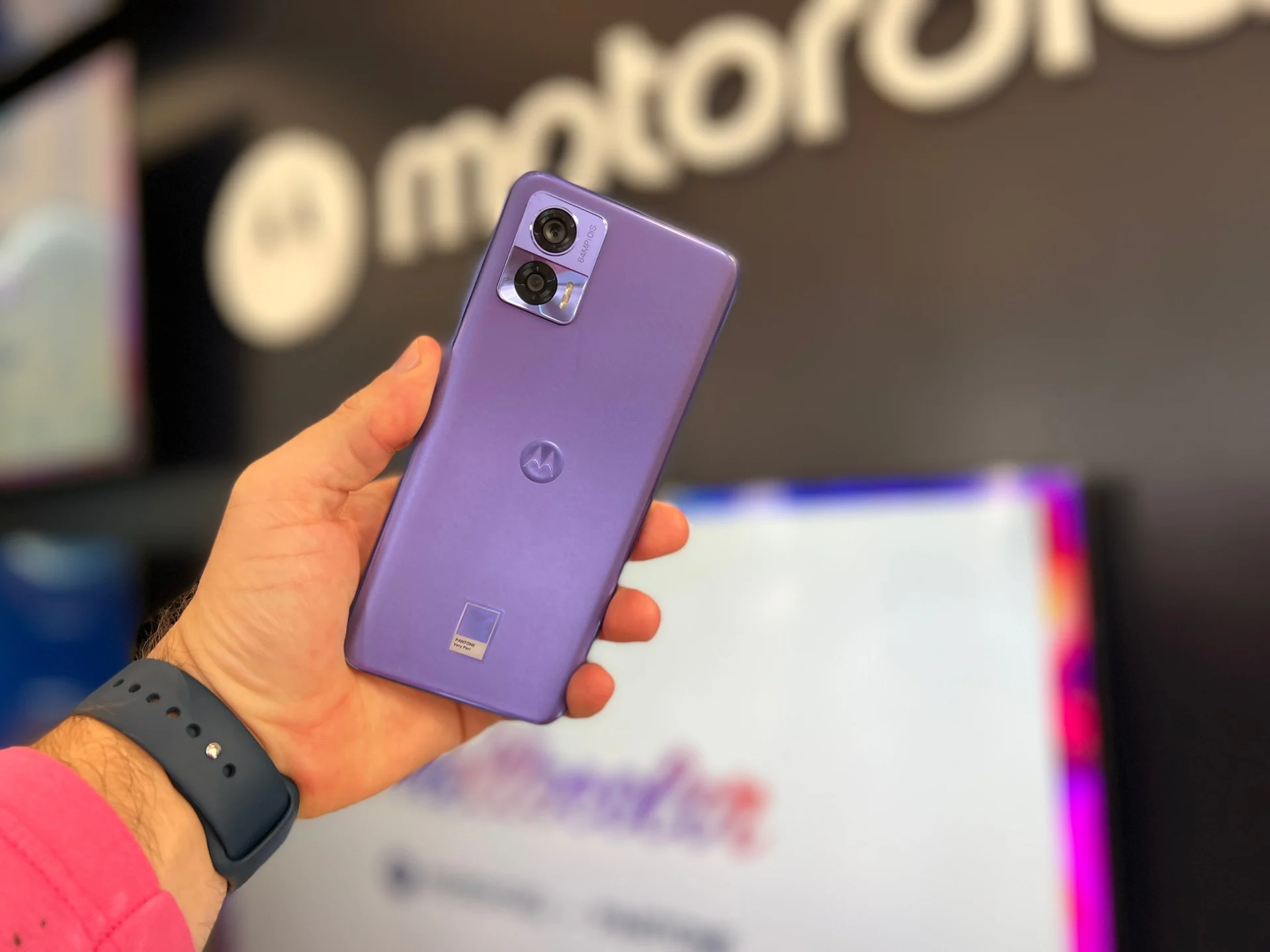 Pessoa segurando um celular Motorola Edge 30 Neo na cor lilás, com logo da Motorola ao fundo