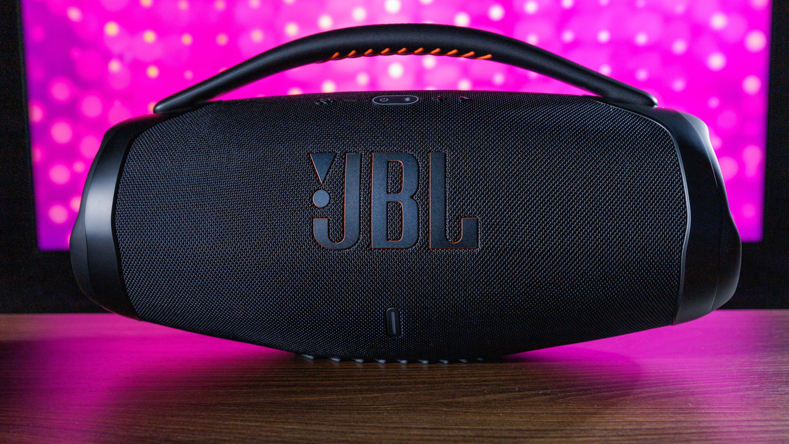 Fundo rosa, destacando a caixa de som preta da marca JB em uma bancada marrom