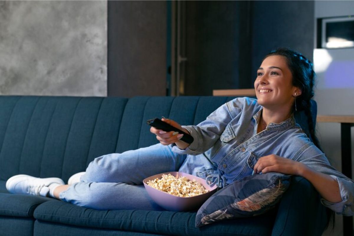 Uma mulher está sentada no sofá enquanto aponta o controle da televisão. Entre suas pernas há um pote de pipoca