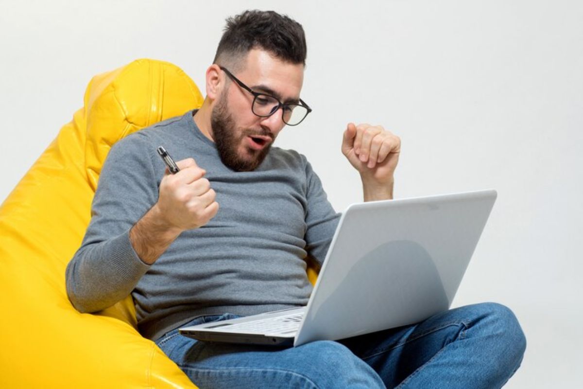 Um homem com o notebook no colo está com as mãos fechadas como quem comemora