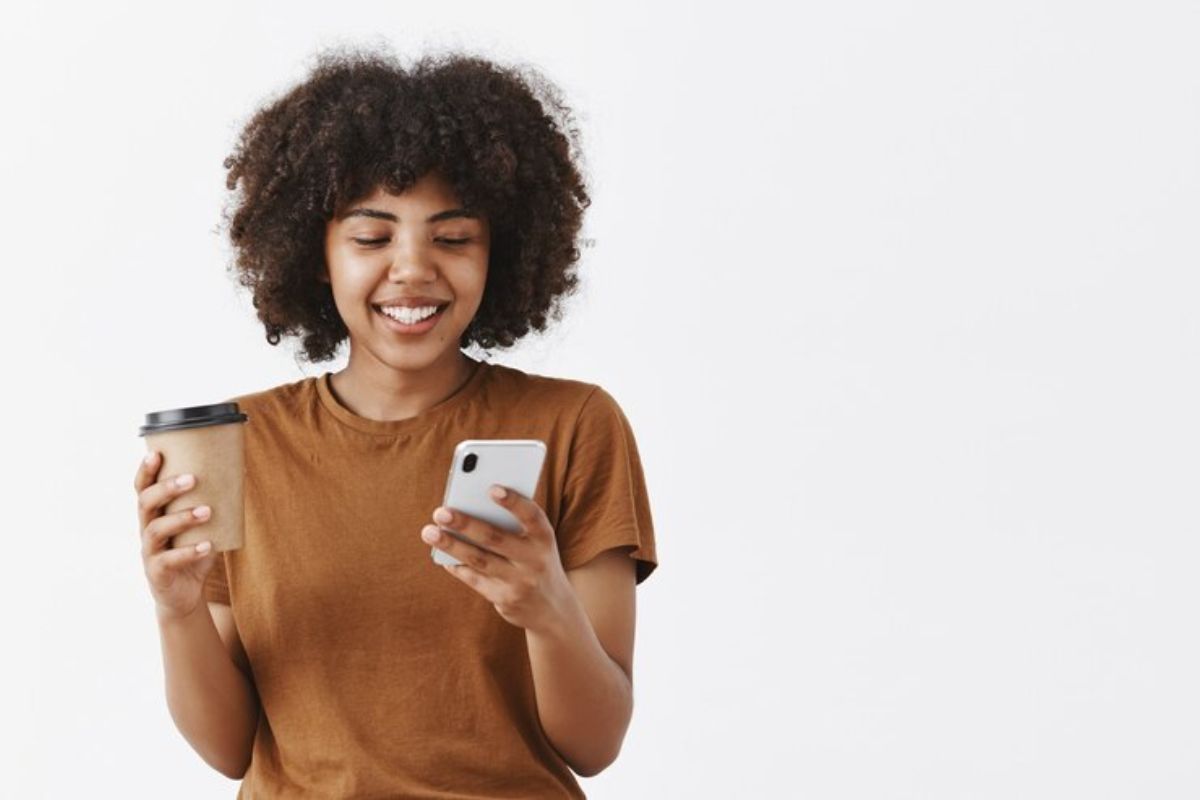 Uma menina de camisa marrom segurando um dos 3 Melhores Celulares Chineses em uma mão e um copo de café na outra