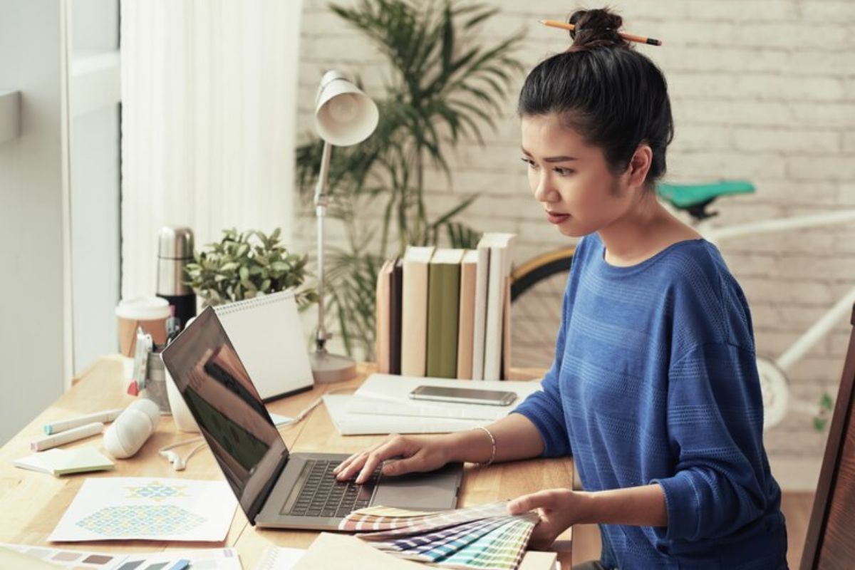 Uma designer sentada, trabalha em seu notebook. Em uma das mãos, ela segura diversas fitas de cores