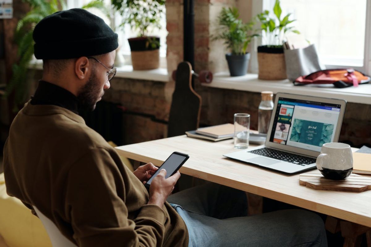 Um homem está sentado no escritório. Ele está sentado em frente ao seu computador e segura o celular na mão usando um dos Melhores Aplicativos para Editar Fotos em 2024