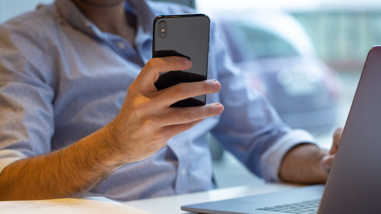 Homem usando celular baratinho enquanto trabalha no notebook