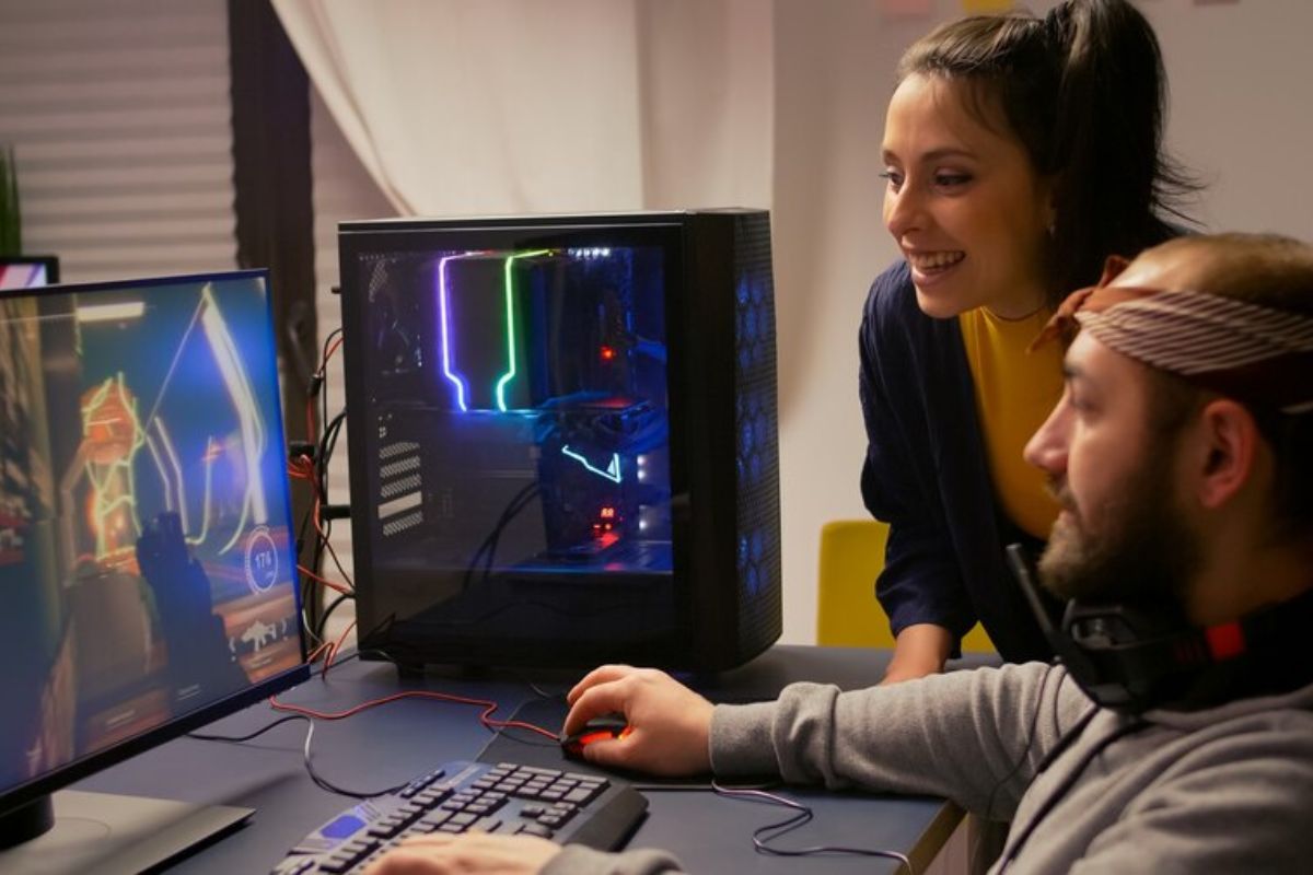 Um casal. O marido está sentado na frente do computador jogando enquanto sua esposa acompanha ao lado agachada