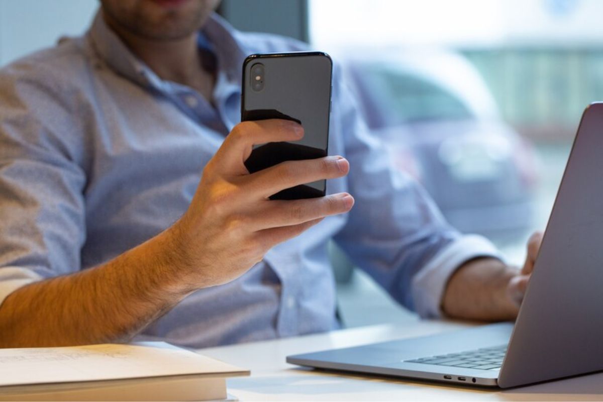 Um homem com camisa social azul sentado mexe em seu celular. Ele está sentado na frente do seu notebook