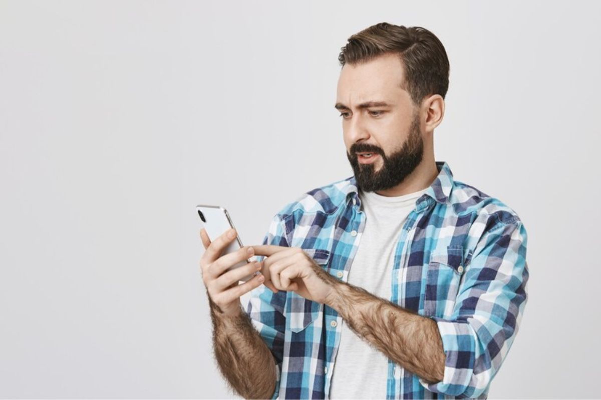 Um homem com camisa xadrez mexe em seu celular branco